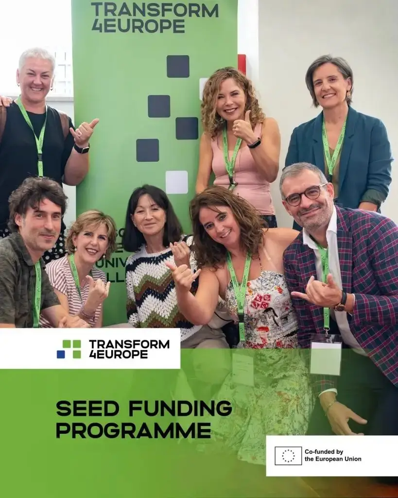 A group of eight smiling adults, wearing conference lanyards, pose together and gesture thumbs up in front of a Transform4Europe banner. Below them, text reads “Seed Funding Programme, Co-funded by the European Union.”.